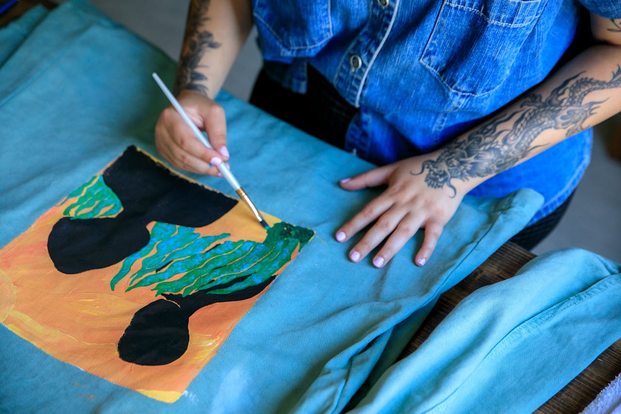 A woman with tattoos paints a colorful design on fabric, showcasing creativity.