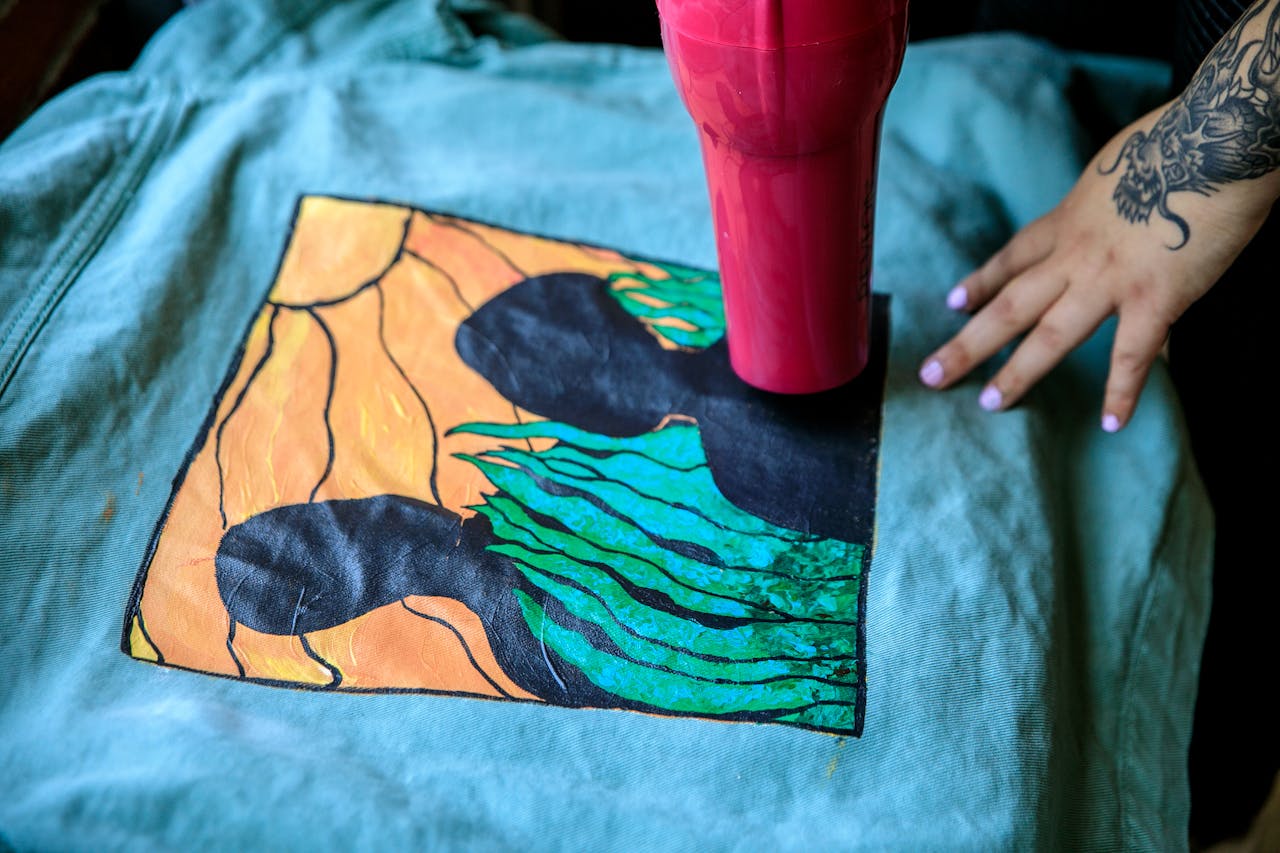 Close-up of a vibrant painted textile being dried with a hairdryer by a tattooed hand.